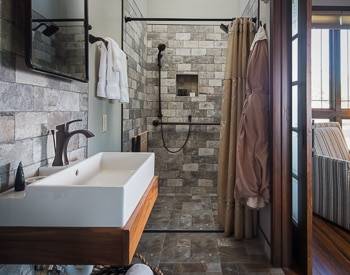 Grey square stone shower stall continuing onto the bathroom floor with a floating white sink, with brown robes hanging next to the shower